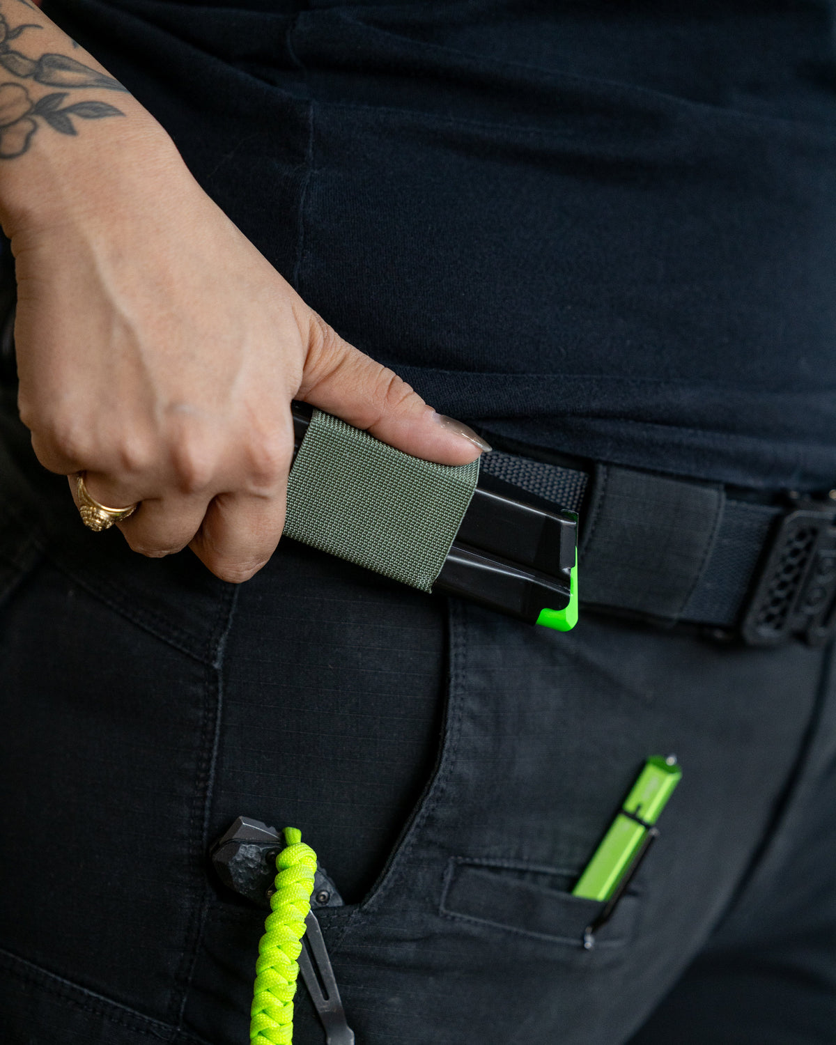 Close-up of a person's hand inserting an olive green rectangular accessory into a black belt holster on dark pants, with a bright neon green cord and matching neon green accessory visible.