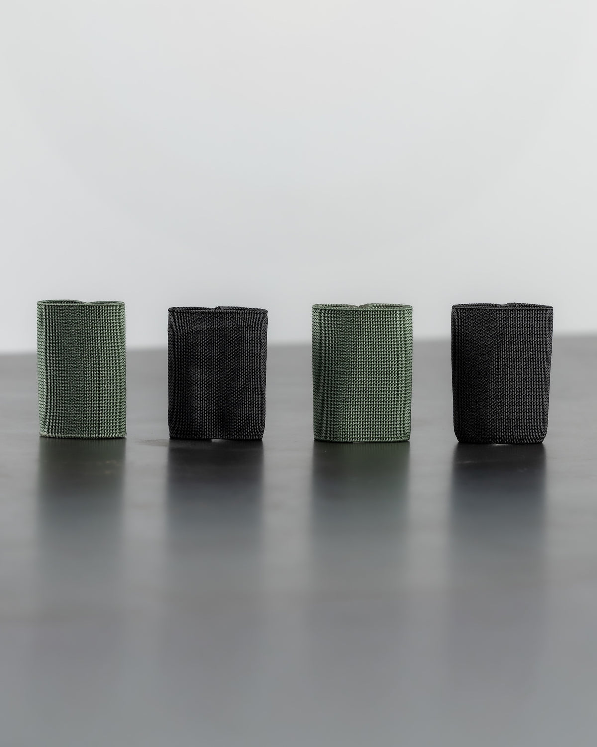 Four rolled SLIM carry bands in alternating green and black colors, arranged in a row on a dark reflective surface against a light gray background.
