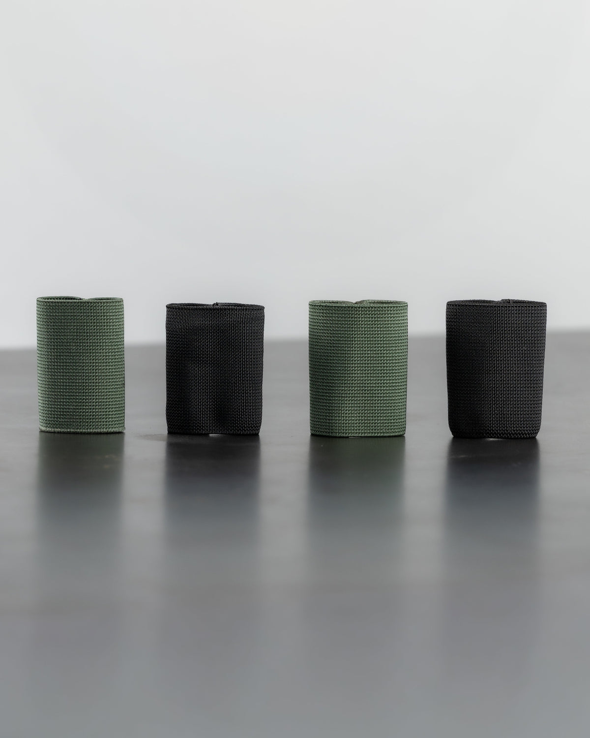 Four textured cylindrical sleeves or sleeves with a ribbed pattern, arranged in a row on a dark reflective surface against a light gray background. Two sleeves are olive green and two are black.